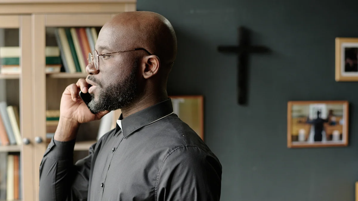 A pastor in a clerical shirt speaking on the phone in front of a wall cross
